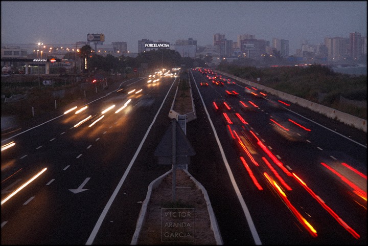 Fotografía de una autovía llena de tráfico con las estelas de los faros encendidos y la ciudad de fondo