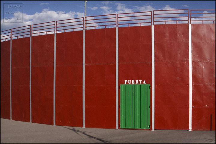Fotografía del exterior de una plaza de toros pintada de rojo con una puerta verde sobre la que aparece escrito "puerta"